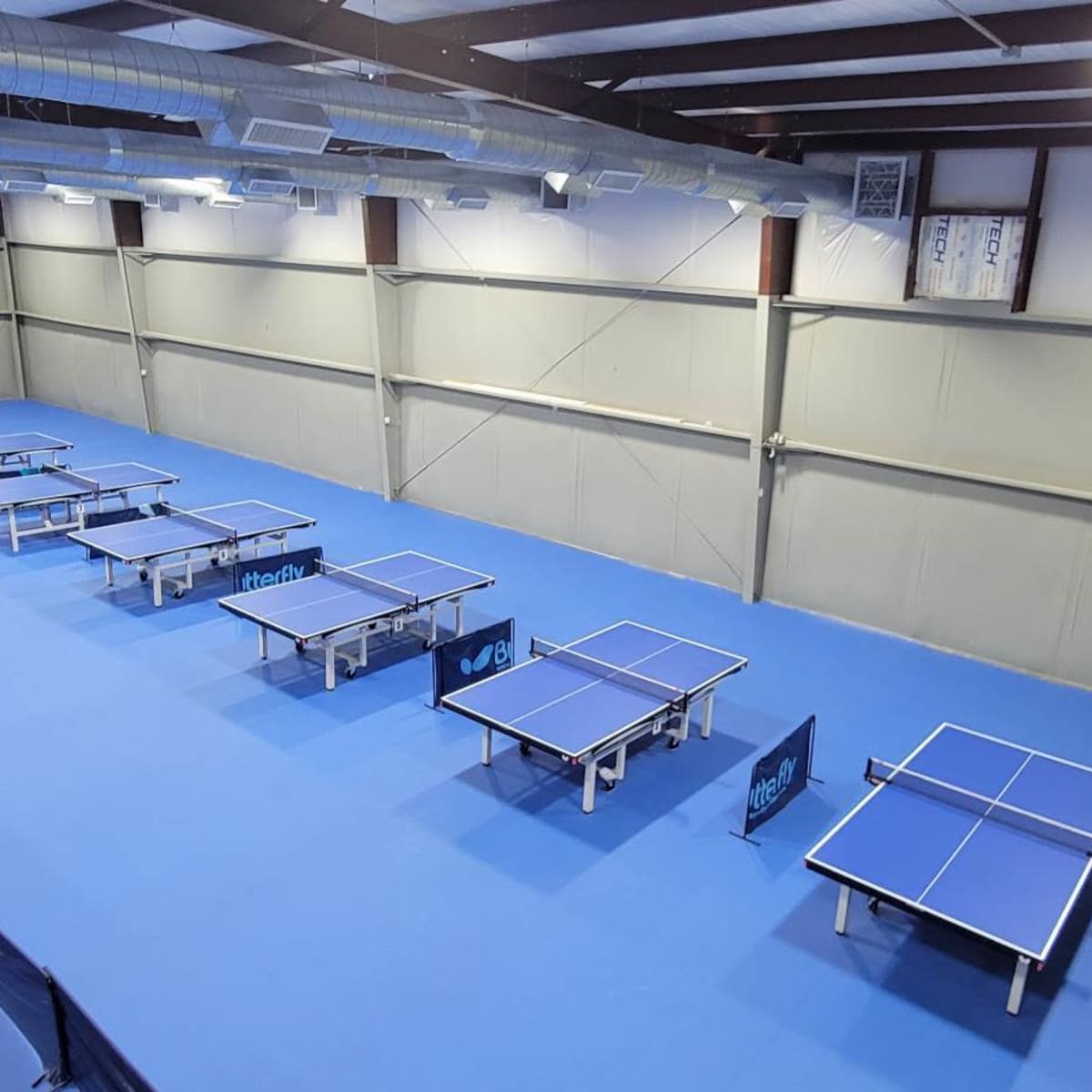 A spacious indoor table tennis facility featuring multiple blue tables arranged in rows with seating along the sides.