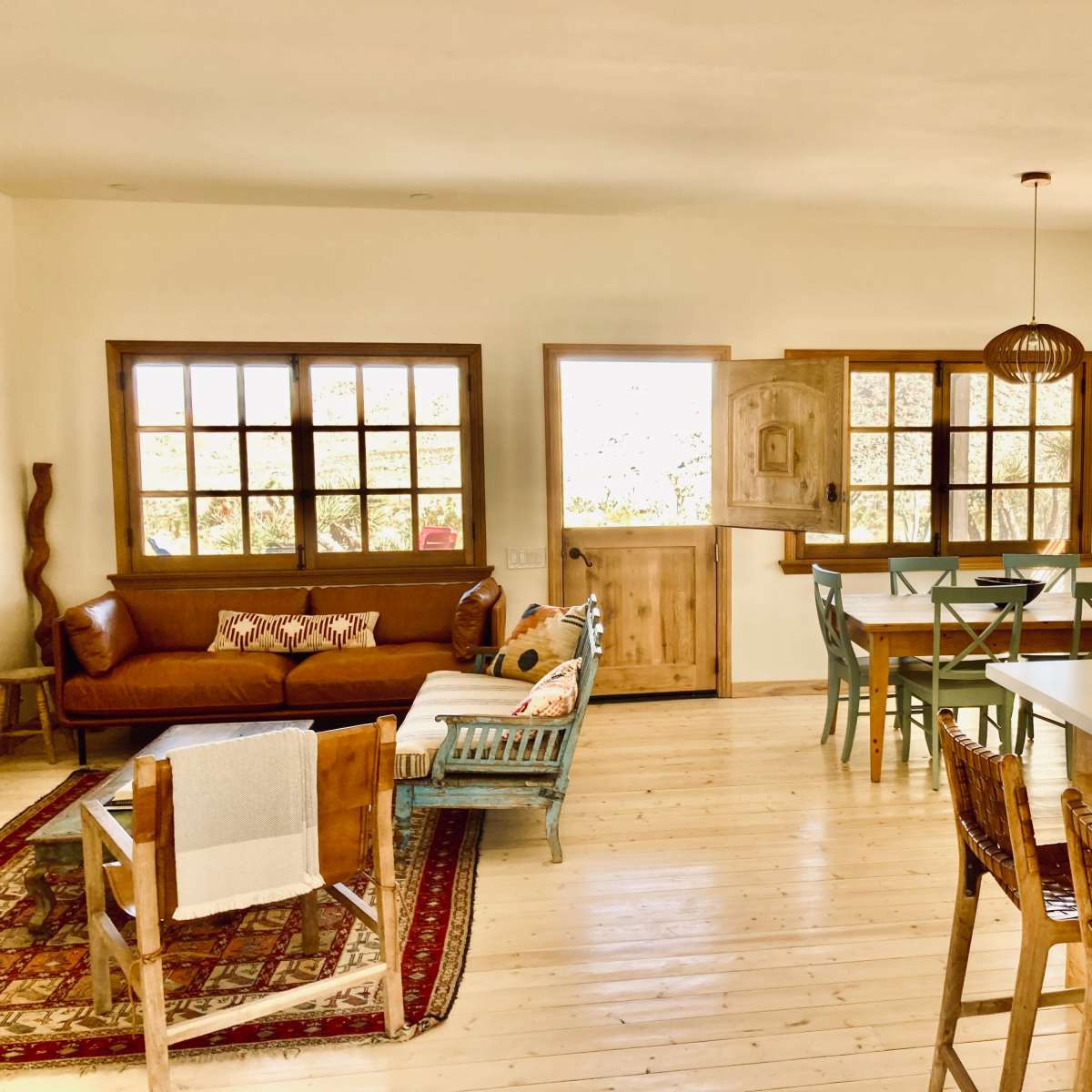 A bright, open-plan living area with a sofa, dining table, and kitchen bar, featuring wooden furniture and large windows.