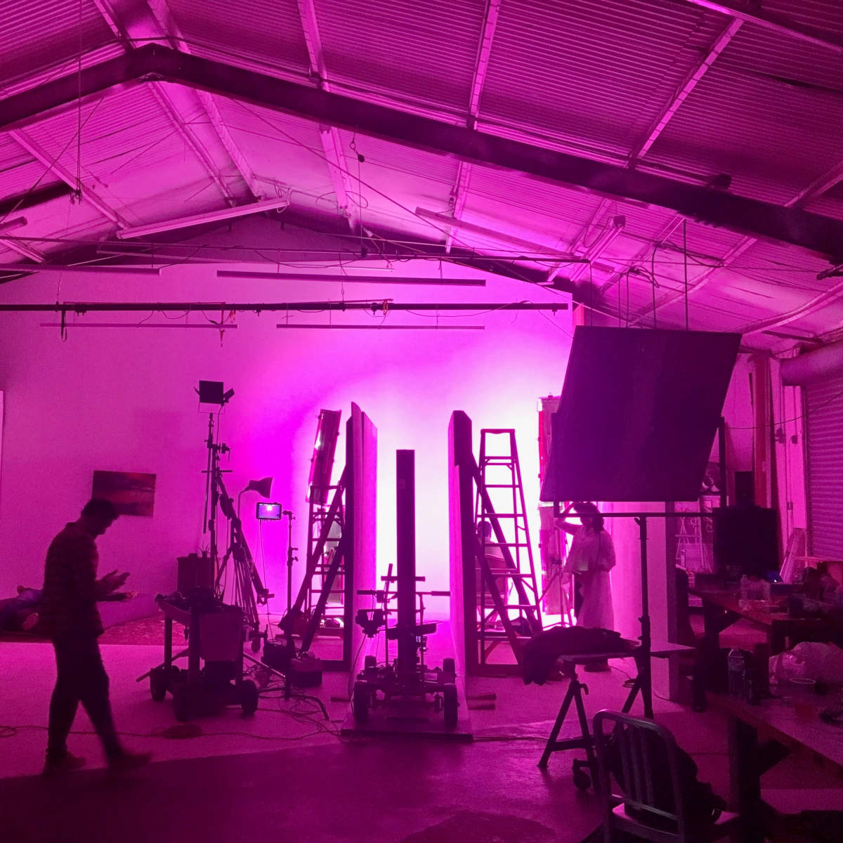 A dimly lit studio space featuring pink lighting, several ladders, equipment setup, and two people moving around.