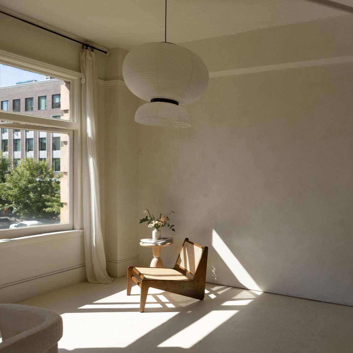 A sunlit room features a simple wooden chair, a small side table with a vase of flowers, and a large mirror reflecting the space.
