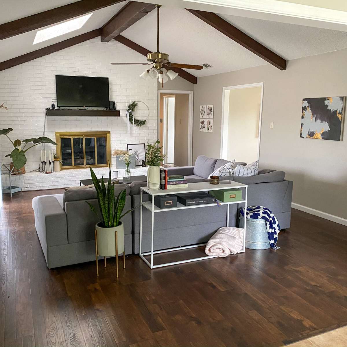 The living room features a gray sectional sofa facing a white-brick fireplace and a television, with wooden beams overhead and various decorative elements throughout.