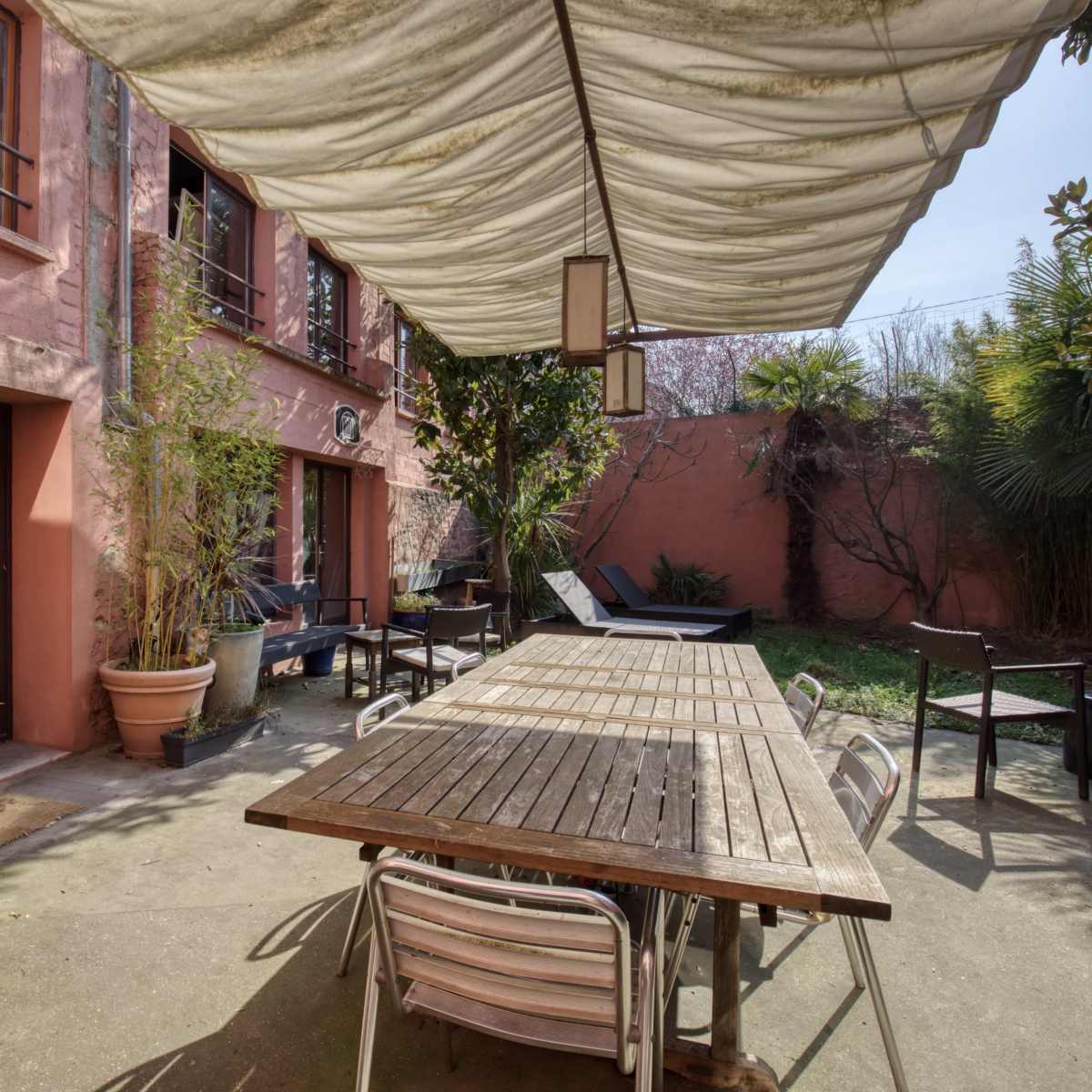 A courtyard with a wooden dining table and chairs beneath a canopy, surrounded by potted plants and buildings with pink walls.