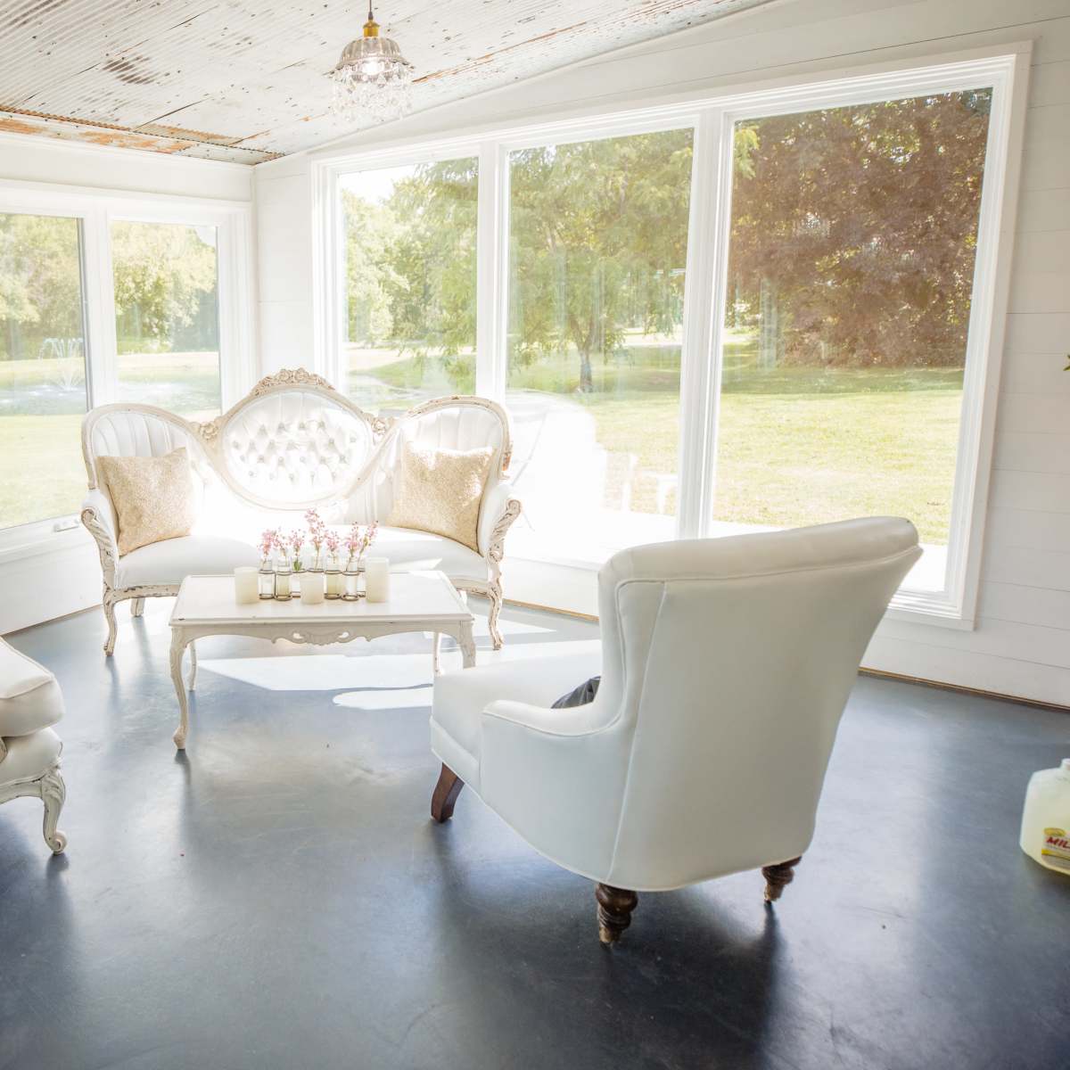 A bright, airy room with large windows, featuring a white upholstered set of chairs and a decorative sofa arranged around a central coffee table.