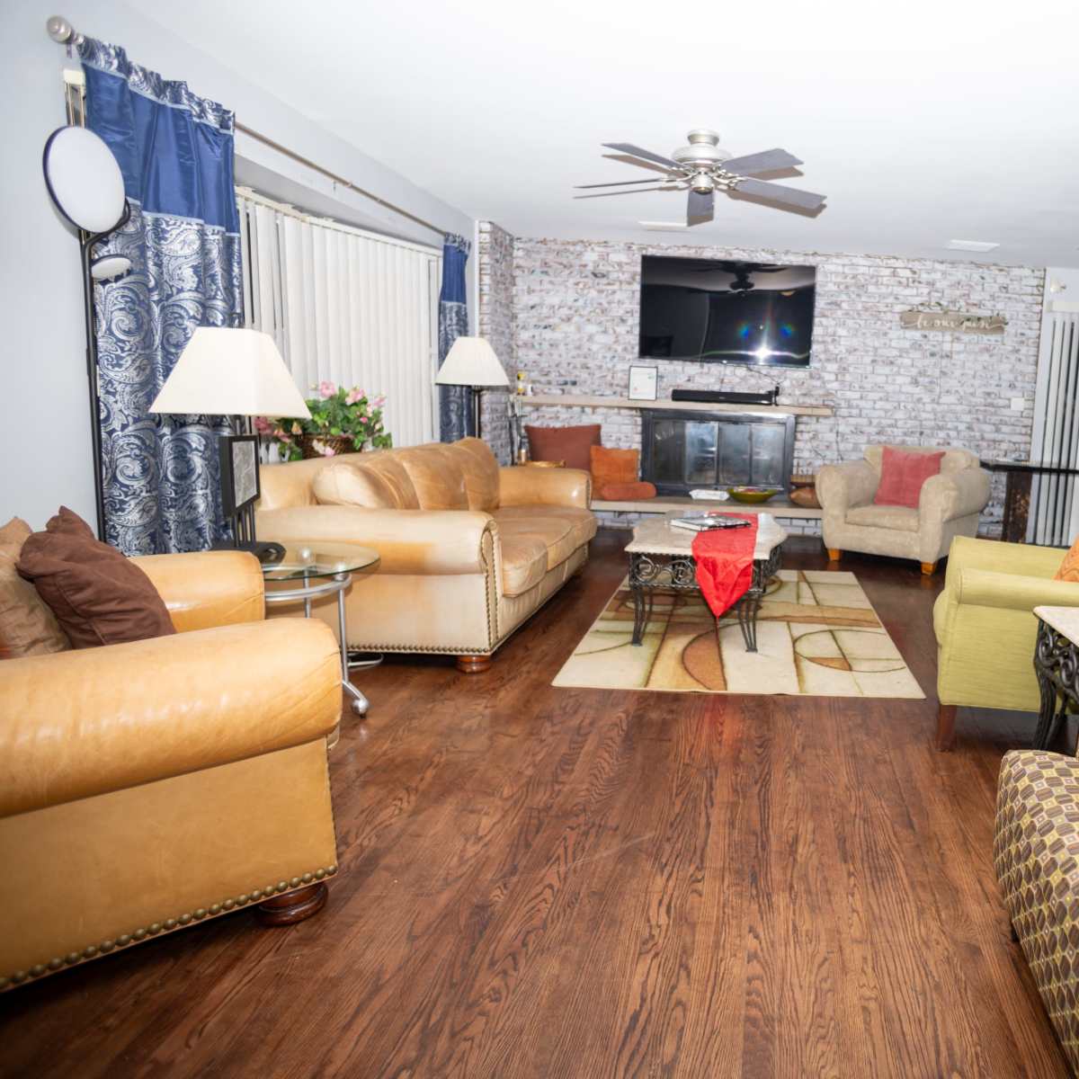 The living room features two leather sofas, an armchair, and a small table, centered around a coffee table on a patterned rug, with a television mounted on a brick wall.