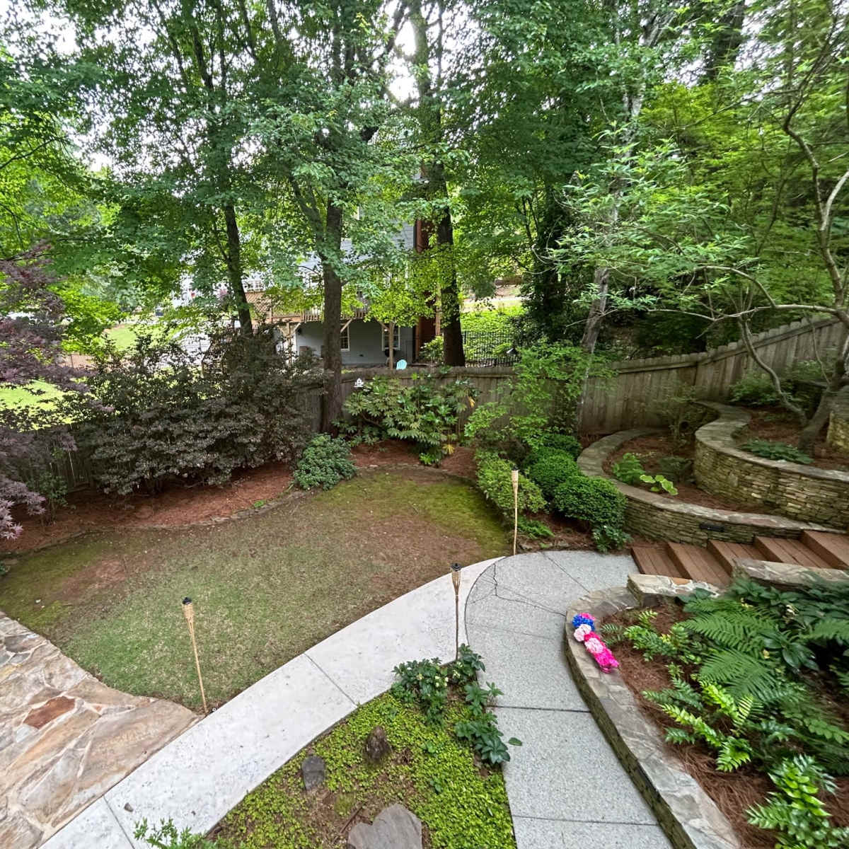 A landscaped backyard featuring stone pathways, patches of grass, trees, and a wooden deck.