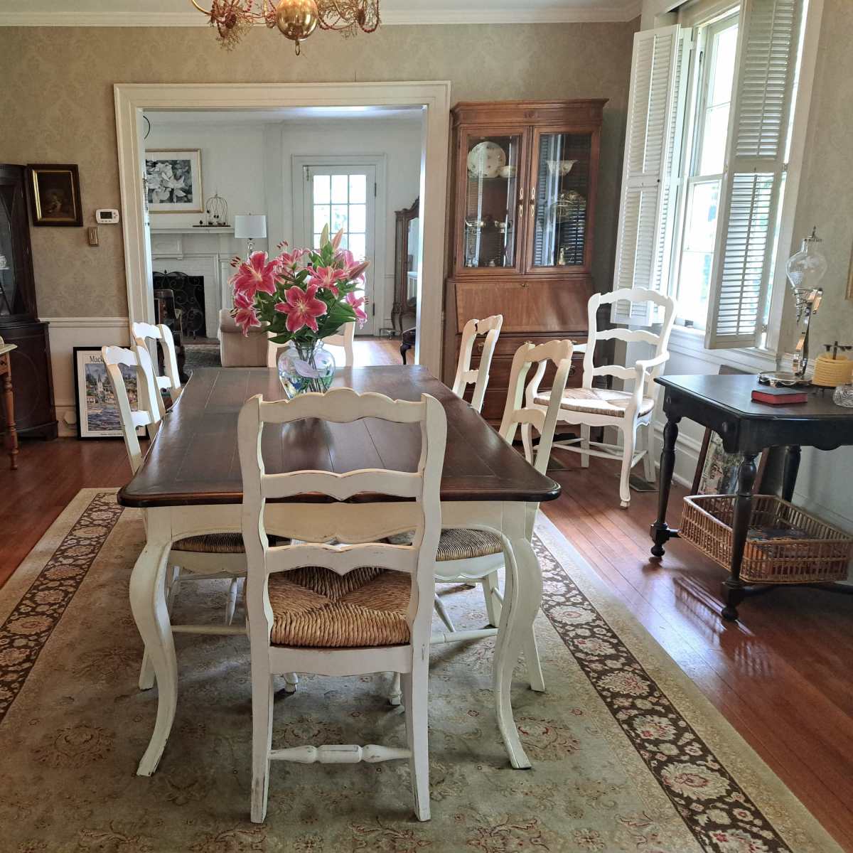 A dining room with a large wooden table surrounded by white chairs, a vase of pink flowers on the table, and a decorative rug on the hardwood floor.