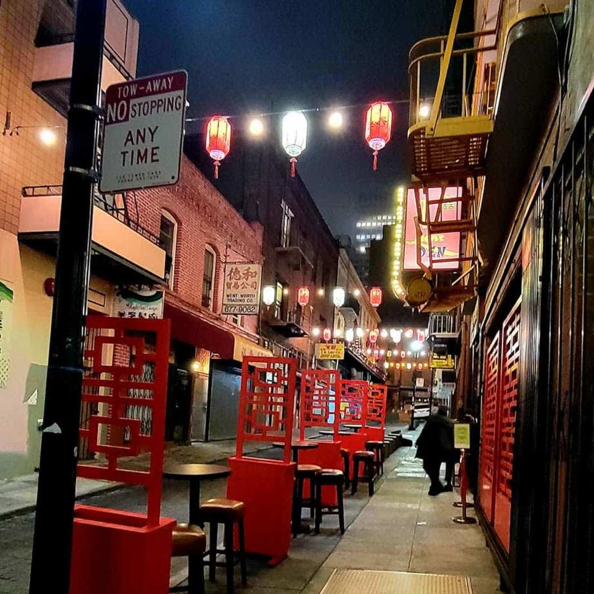 A deserted street in Chinatown features red tables and lanterns illuminated against a night sky.