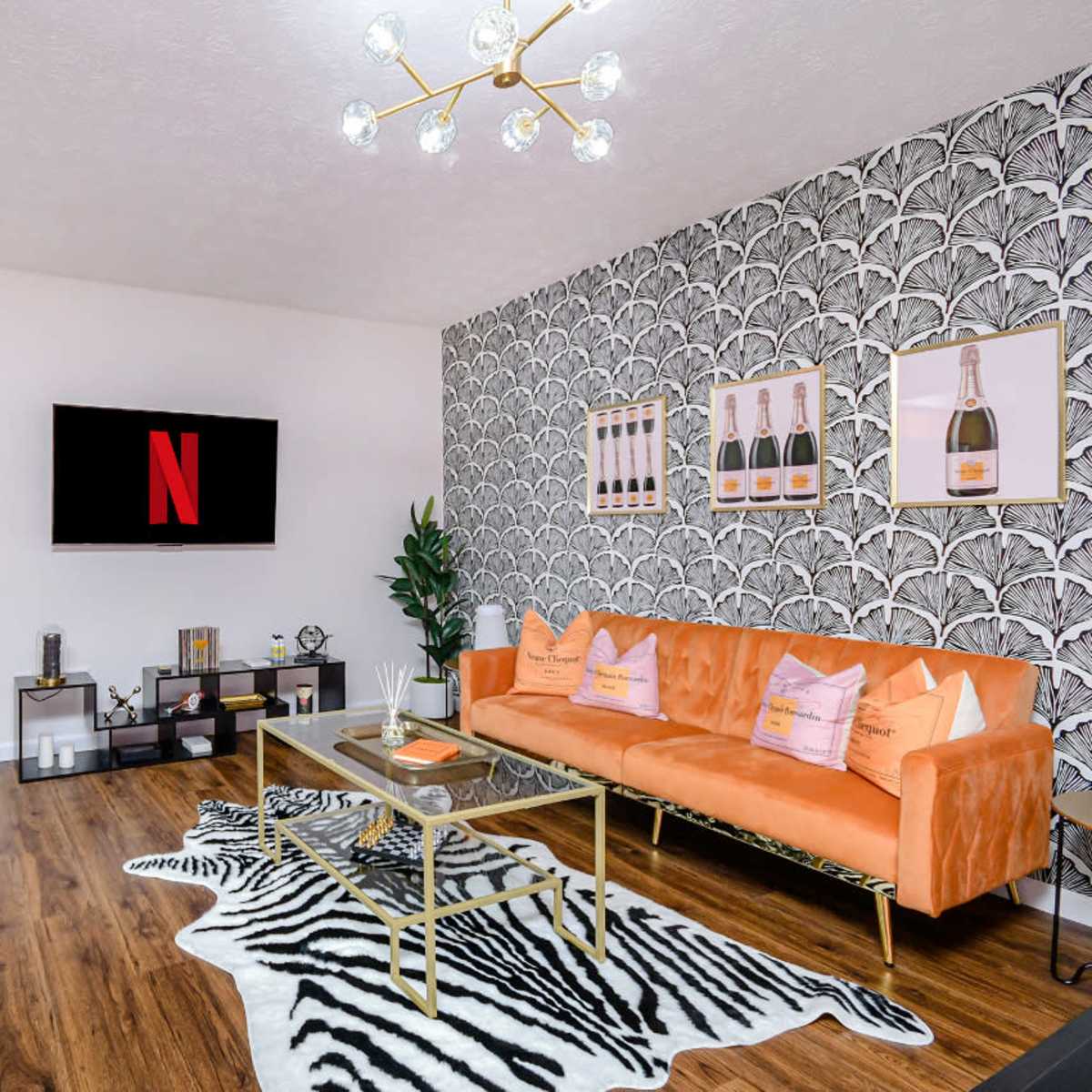 A modern living room featuring a curved orange couch, a glass coffee table on a zebra-patterned rug, and a wall adorned with champagne-themed artwork.