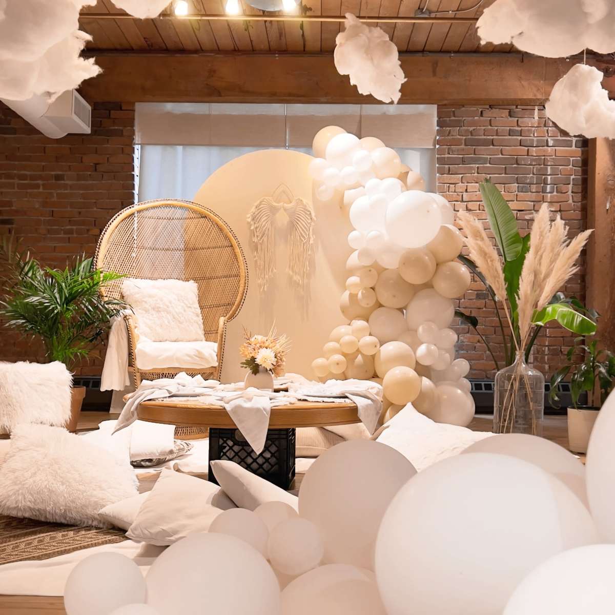 A decorated interior space features a cozy seating area with a rattan chair, a backdrop of balloons, and white cloud-like decorations hanging from the ceiling.
