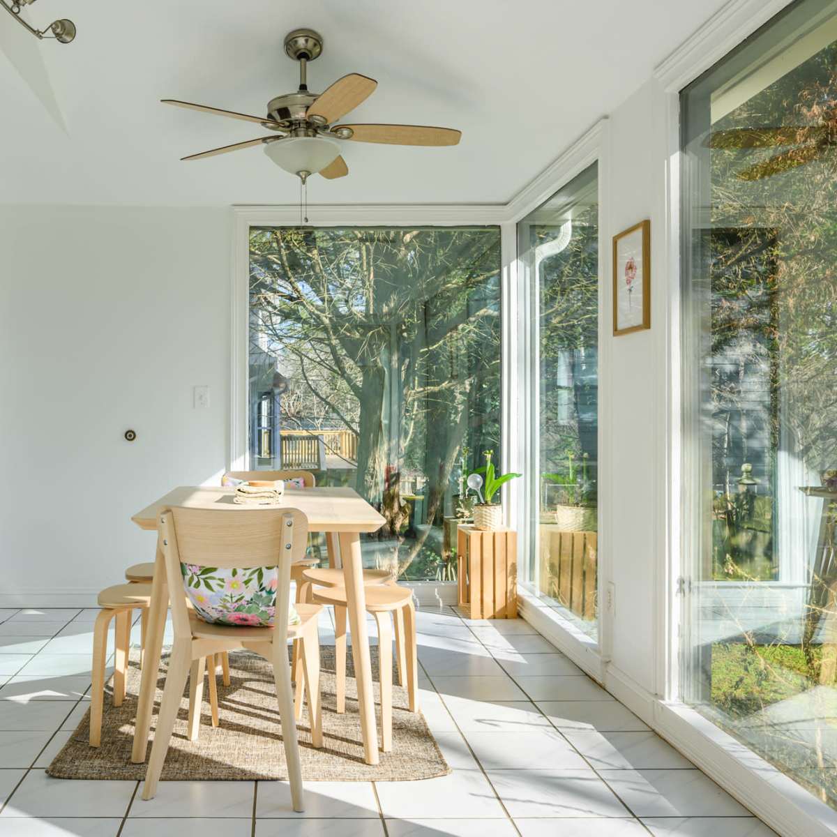 A bright dining area features a table with four chairs near large windows surrounded by greenery.