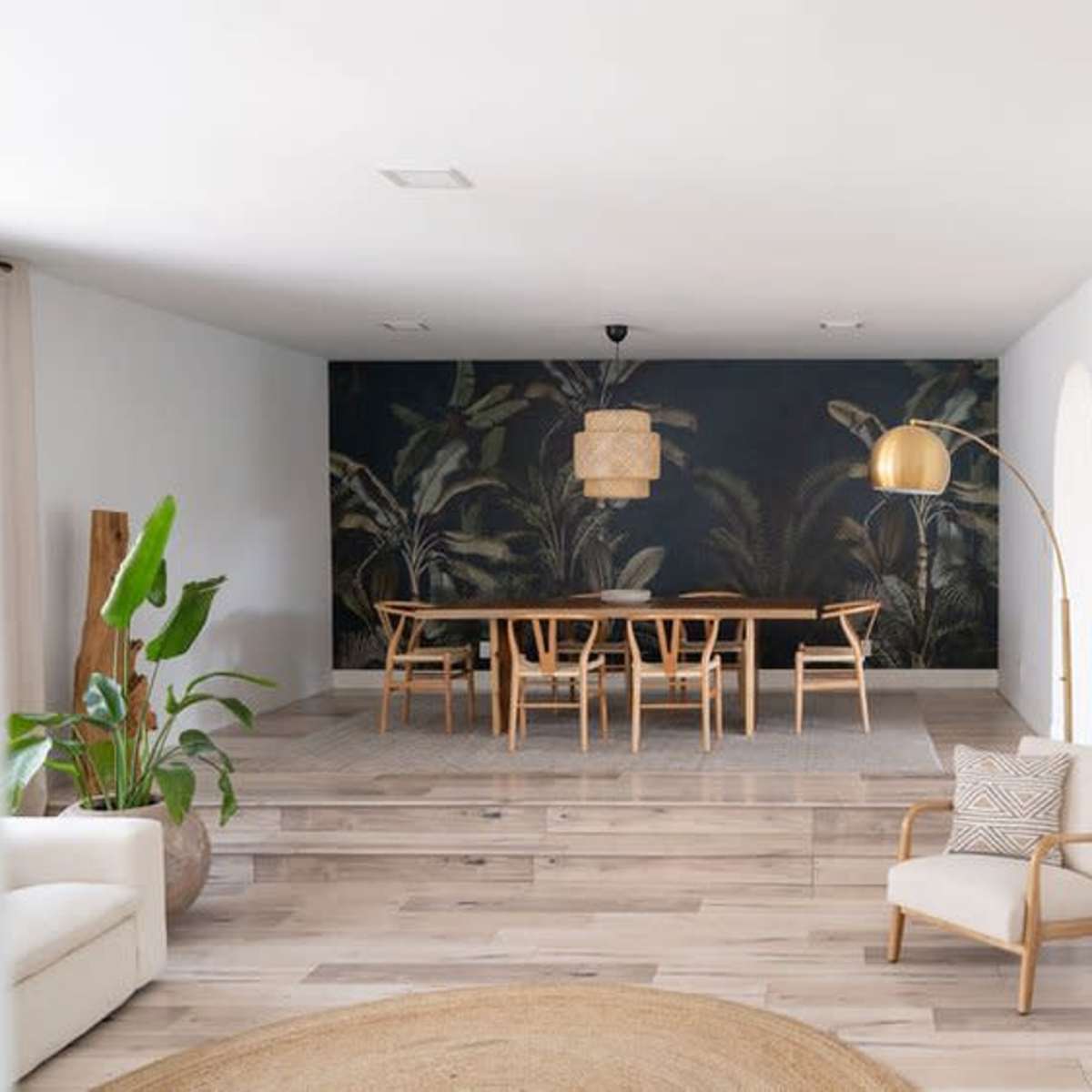 A bright, modern interior featuring a dining area with a wooden table, chairs, and a wall decorated with a tropical mural, along with wooden flooring and a round mirror on the wall.