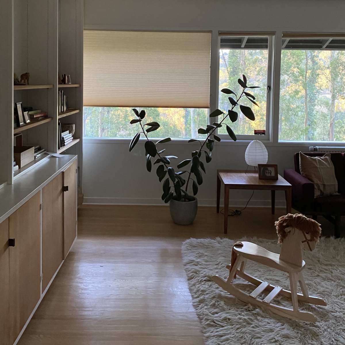 A cozy living room with a large plant in a pot, a wooden rocking horse, a side table, and sunlight filtering through the blinds onto the hardwood floor.