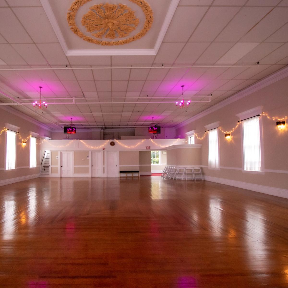 An empty event hall with polished wooden floors, pink lights, and string lights hanging along the walls.