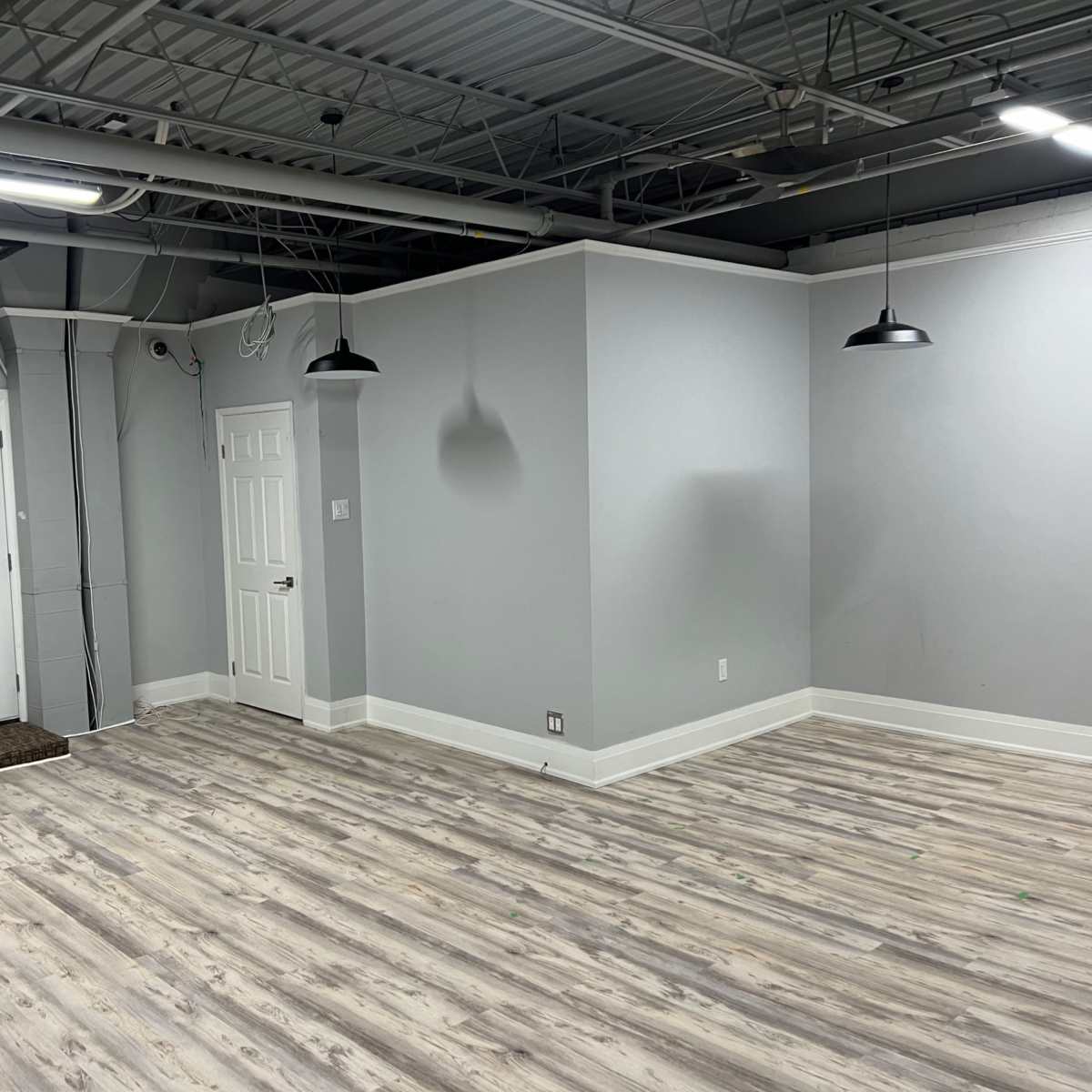 A partially empty, light gray room with two doors, hanging lights, and wooden flooring.