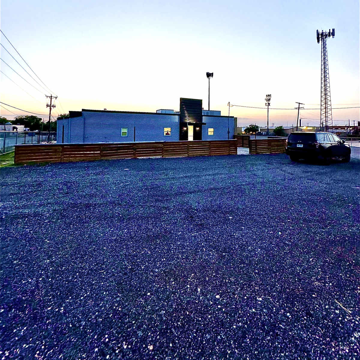 A gray building surrounded by a gravel parking lot, with a cell tower in the background and a clear sky at dusk.