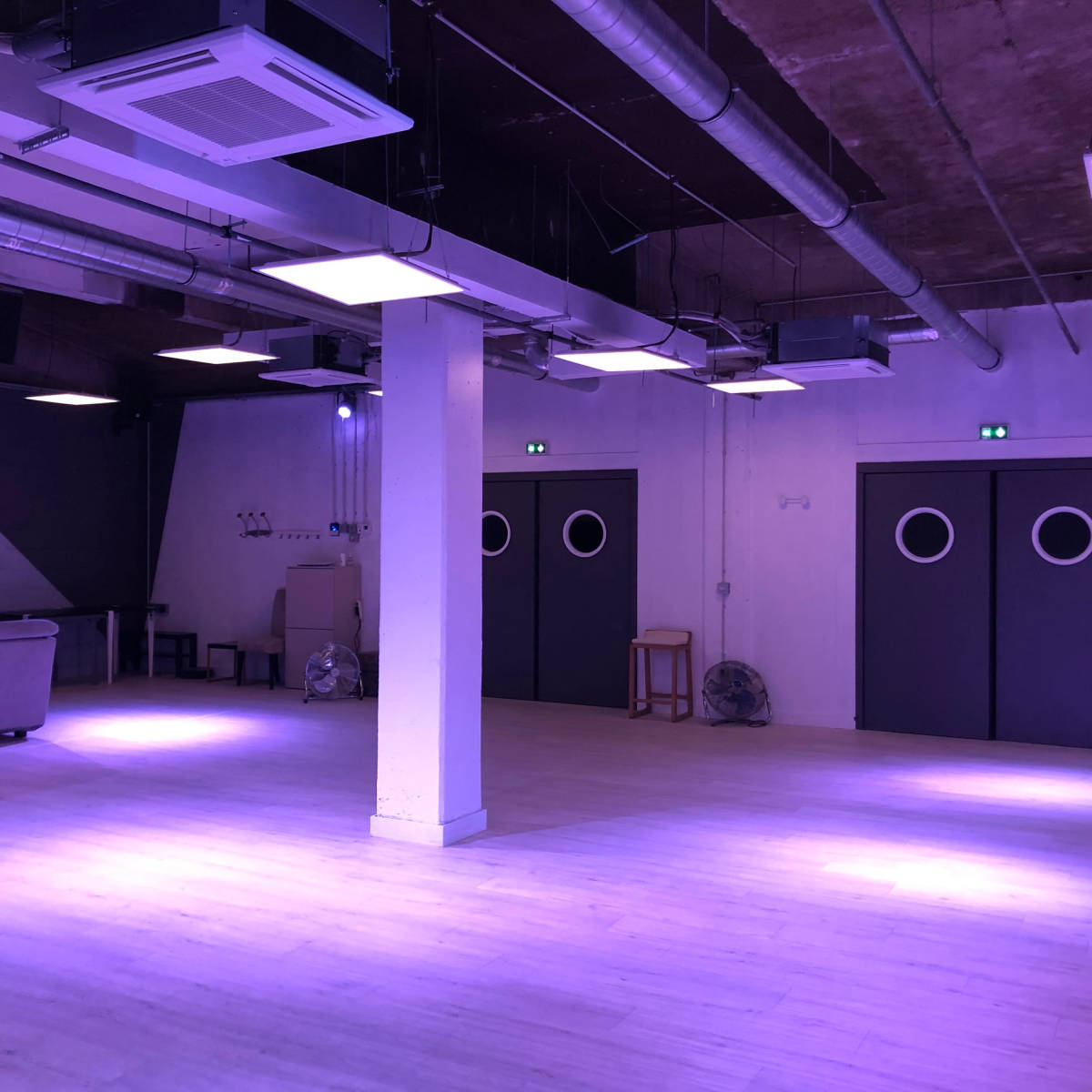 An empty room with wooden flooring, industrial-style ceiling vents, and two closed doors featuring round windows.