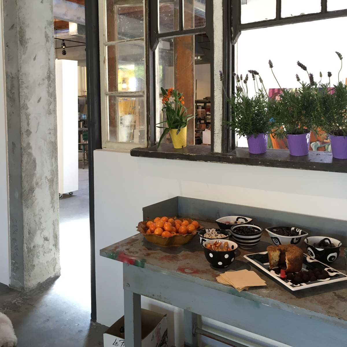A table with fruit, baked goods, and snacks is set against a backdrop of large windows and potted plants in a modern space.