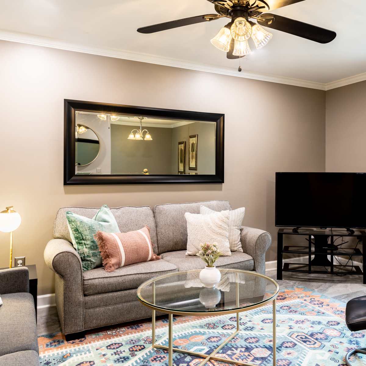 A living room features a gray sofa, a glass coffee table, a black television on a stand, and large windows with double doors leading outside.