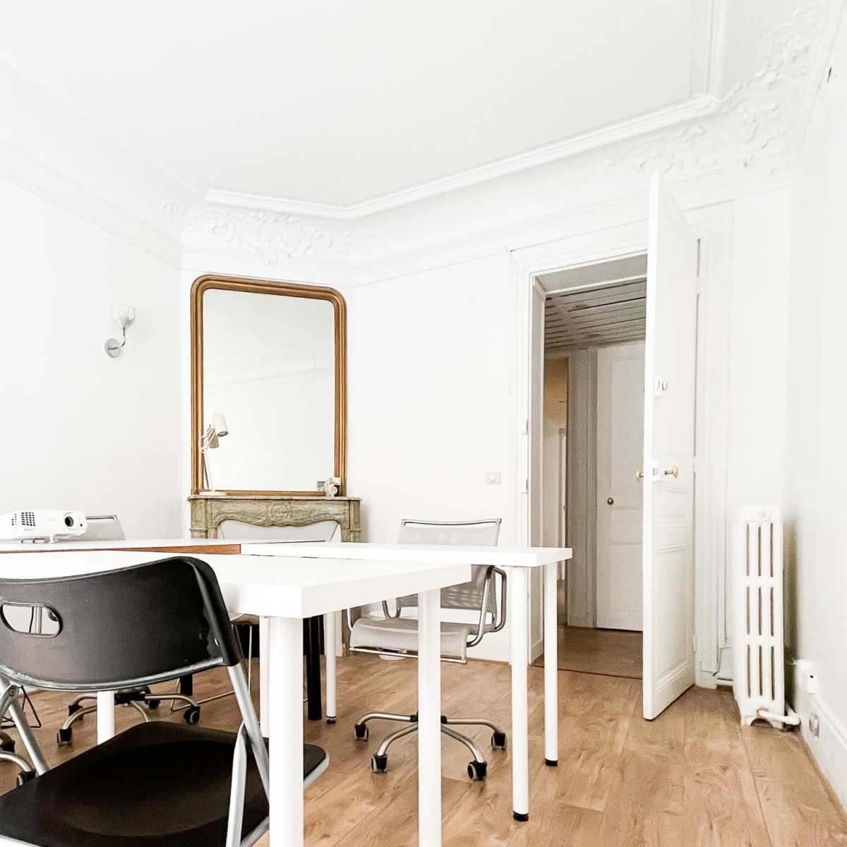 A minimalist office space featuring a white table, several black chairs, a large mirror, and an open door leading to another room.