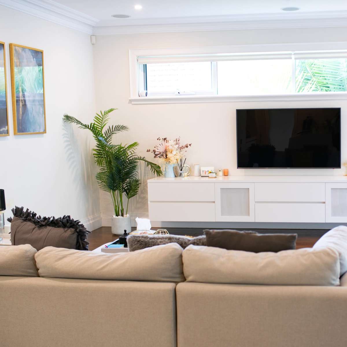 A modern living room features a large television mounted on a white cabinet, with a sofa facing it and indoor plants in the corner.
