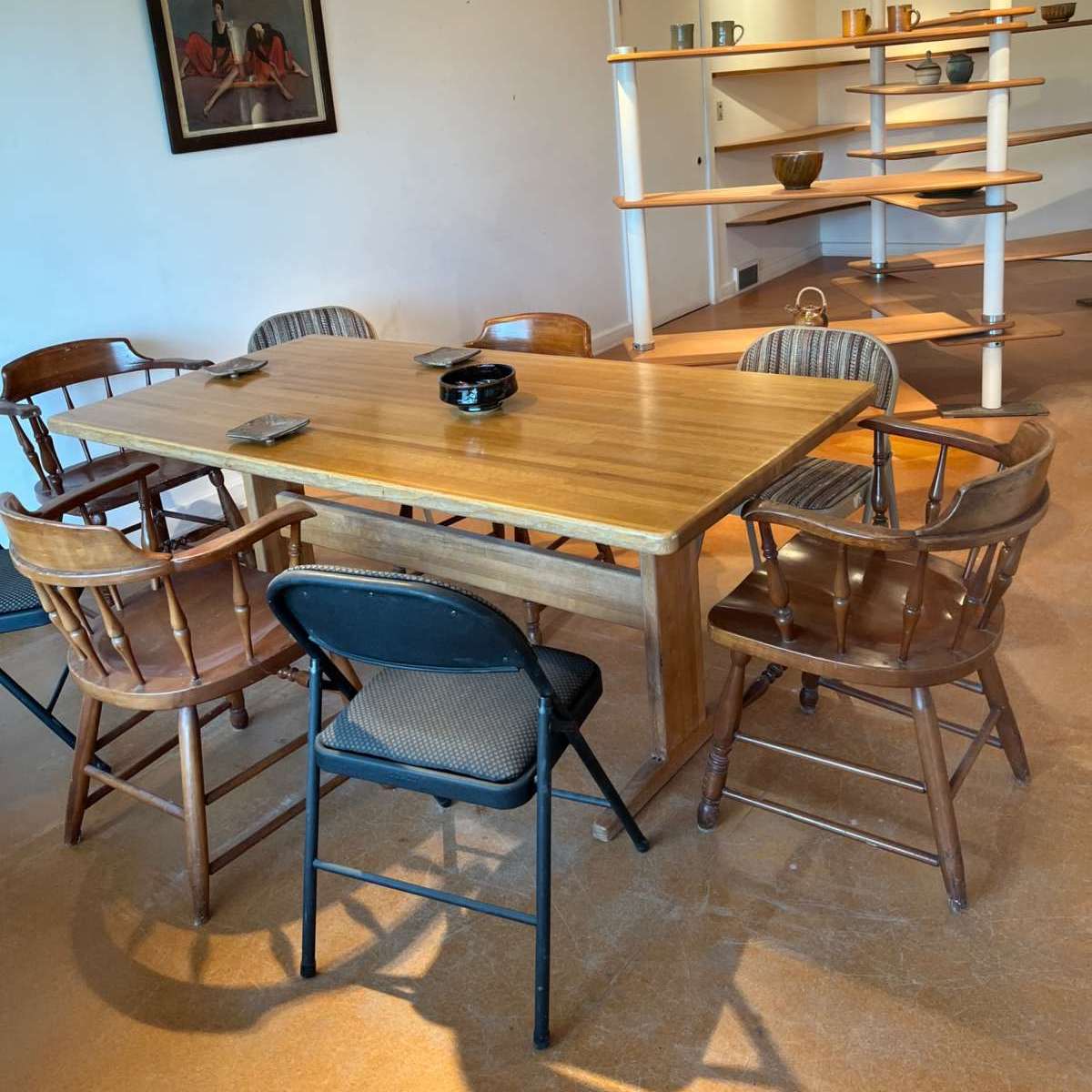 A wooden dining table surrounded by several chairs, with a painting on the wall and a shelf displaying items in the background.