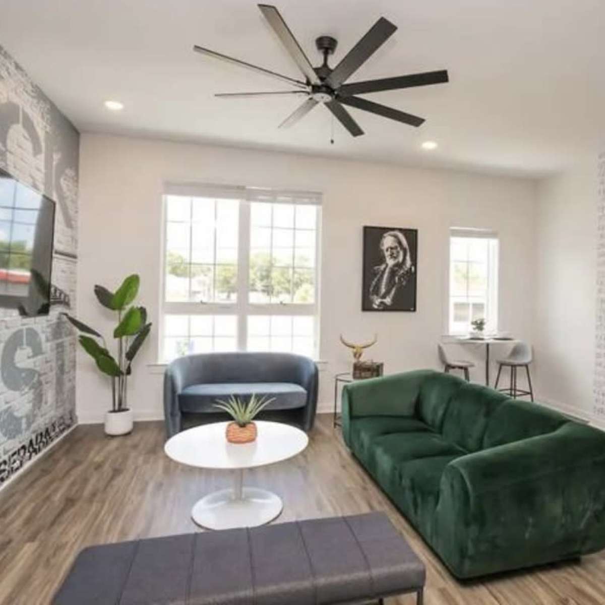 The living room features a green sofa, blue chair, a round coffee table with a plant, a large wall-mounted TV, and large windows allowing natural light.