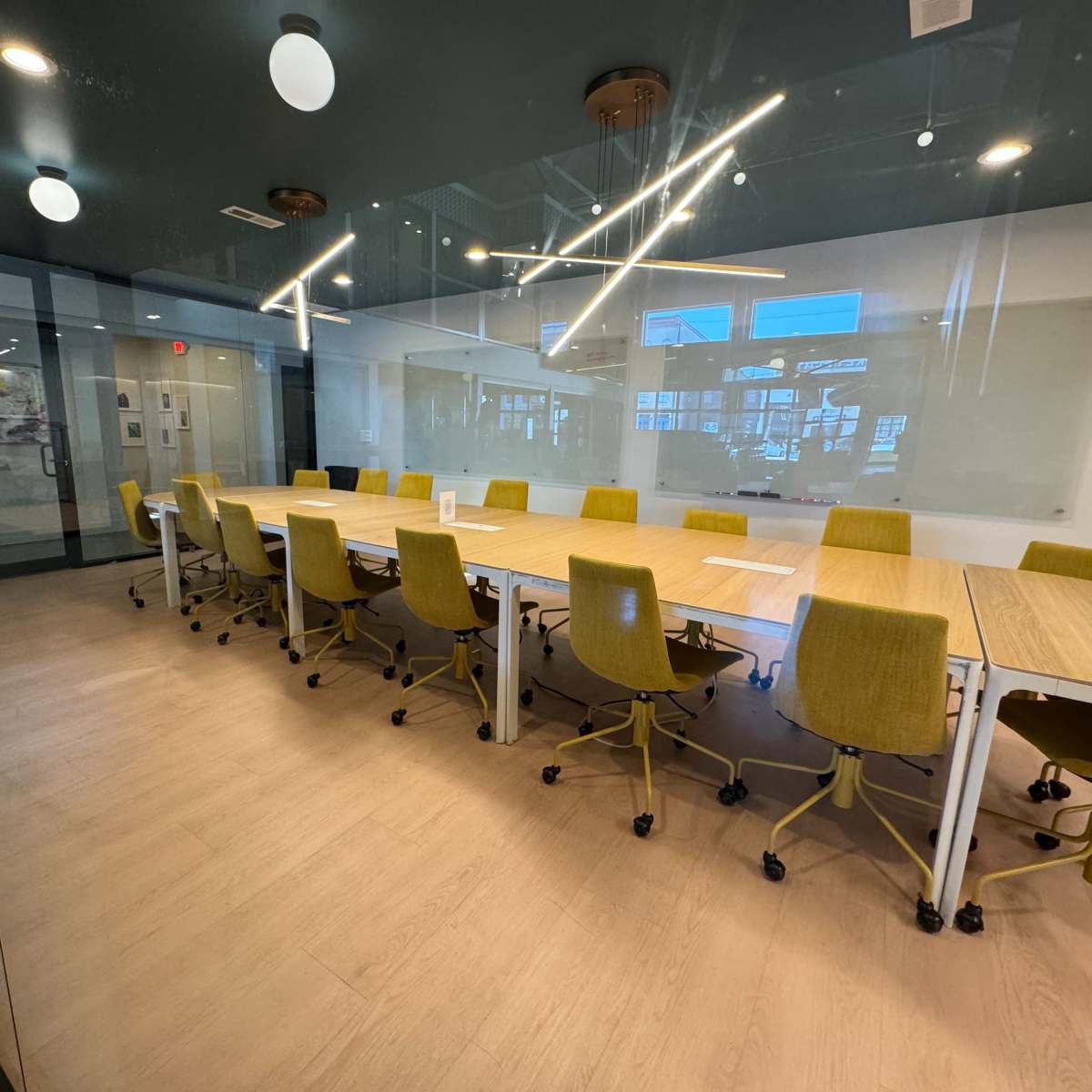 A large, modern conference room features a long wooden table surrounded by yellow office chairs, with large windows allowing natural light to fill the space.