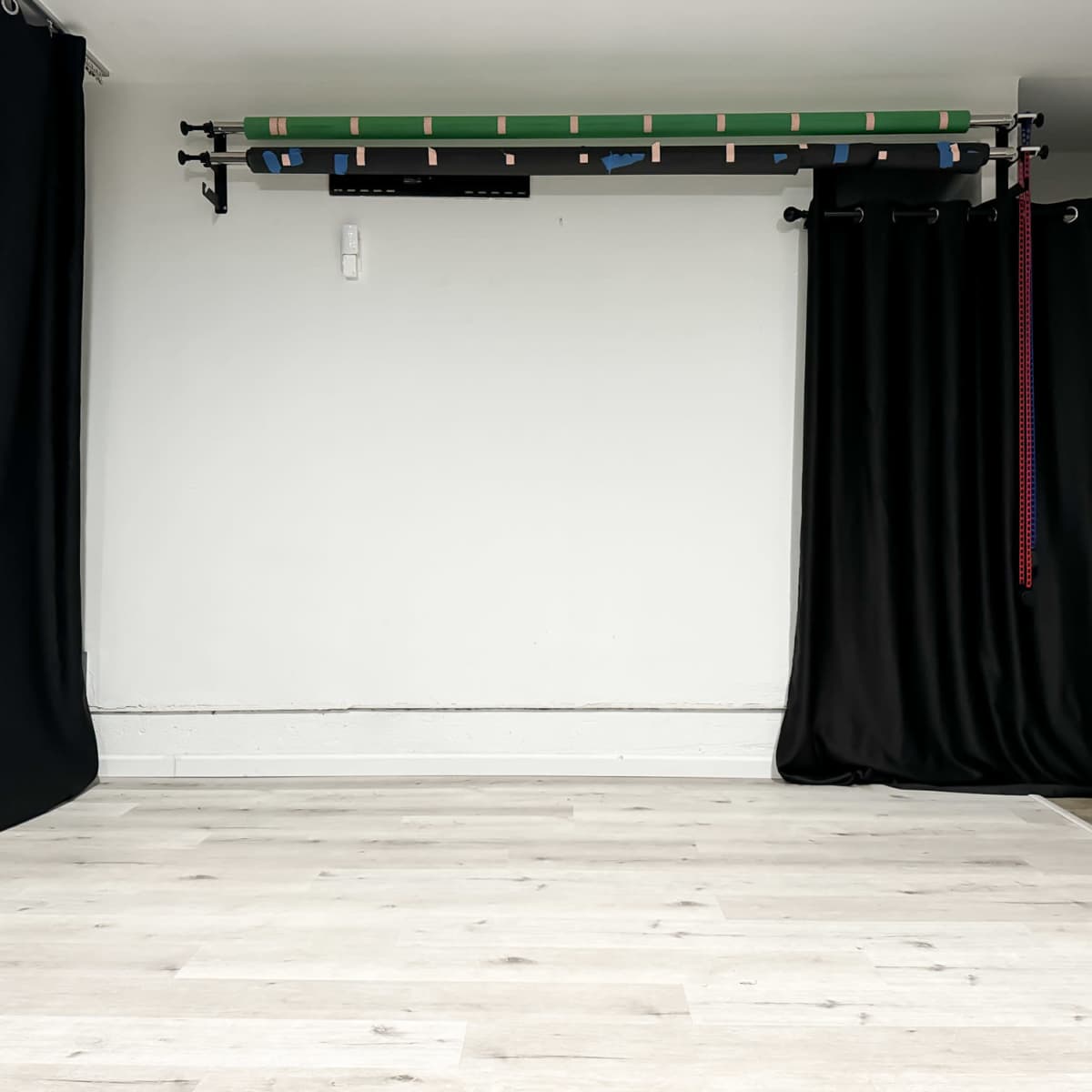 An empty room with light wooden flooring, a plain white wall, and black curtains on one side, along with equipment mounted on the wall above.