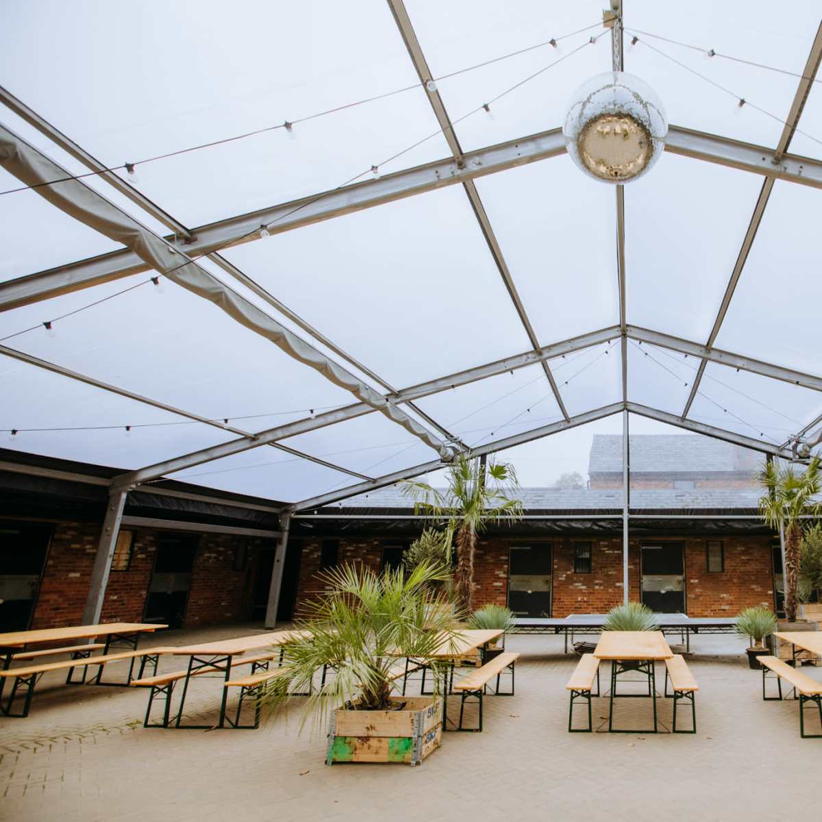 An open-air venue features metal framework and wooden tables arranged under a large canopy with a mirrored disco ball hanging from the center.