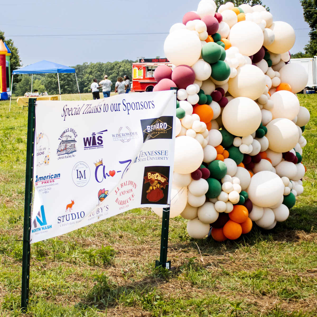A colorful balloon arrangement stands beside a sign thanking sponsors at an outdoor event.