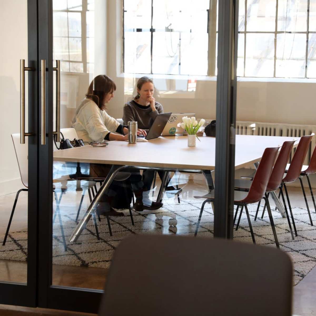 Two women are sitting at a table in a well-lit office, working on laptops with a vase of flowers in the center.