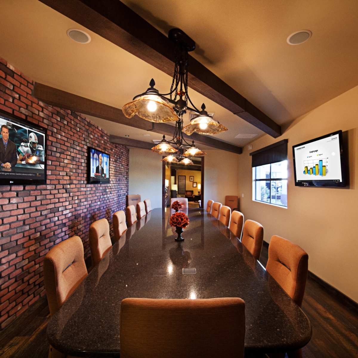 A conference room with a long table, bordered by brick walls and equipped with televisions displaying presentations.