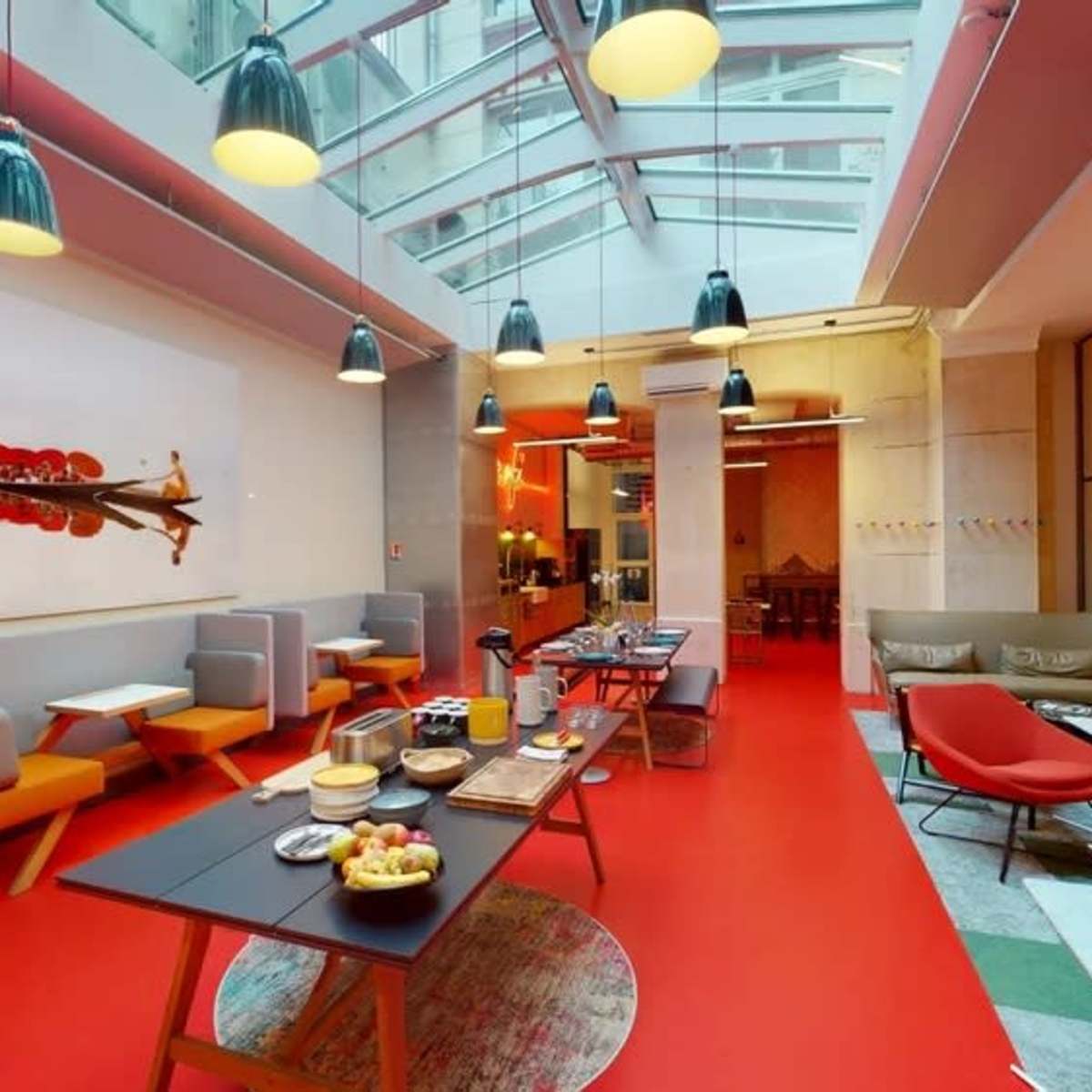 A modern open space with red flooring, large windows, and seating areas featuring gray and orange furniture along with a dining setup.