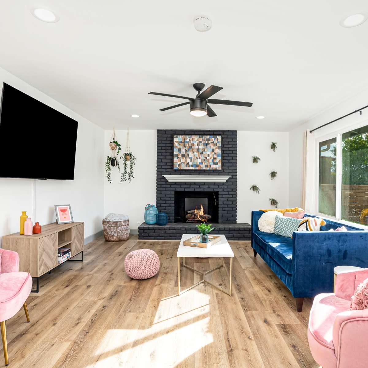 A modern living room features a blue sofa, pink armchairs, a fireplace with a black stone surround, a wall-mounted TV, and large windows allowing natural light to enter.