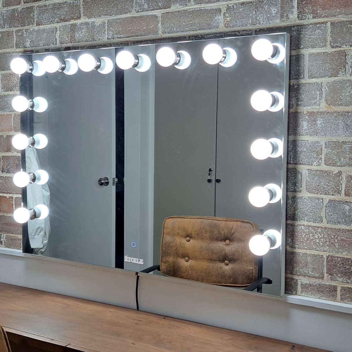A large mirror framed by bright bulbs is mounted on a wall of exposed brick, with a wooden vanity and an upholstered chair in front, and clothing hung on a rack nearby.