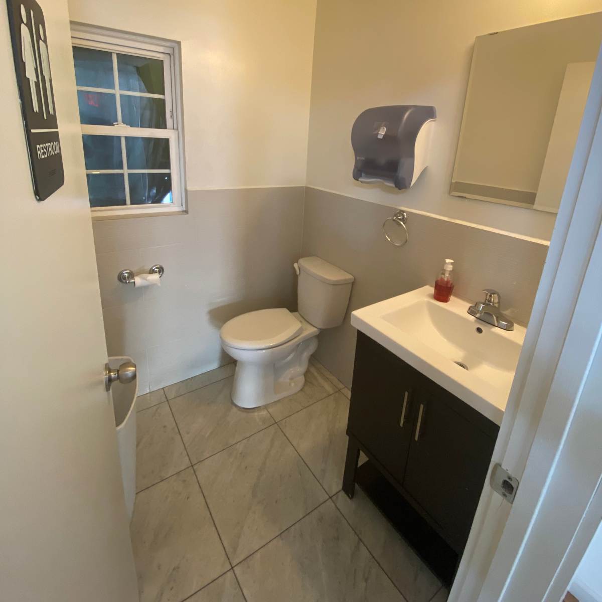 A small restroom featuring a toilet, sink with a cabinet, a mirror, and a window.
