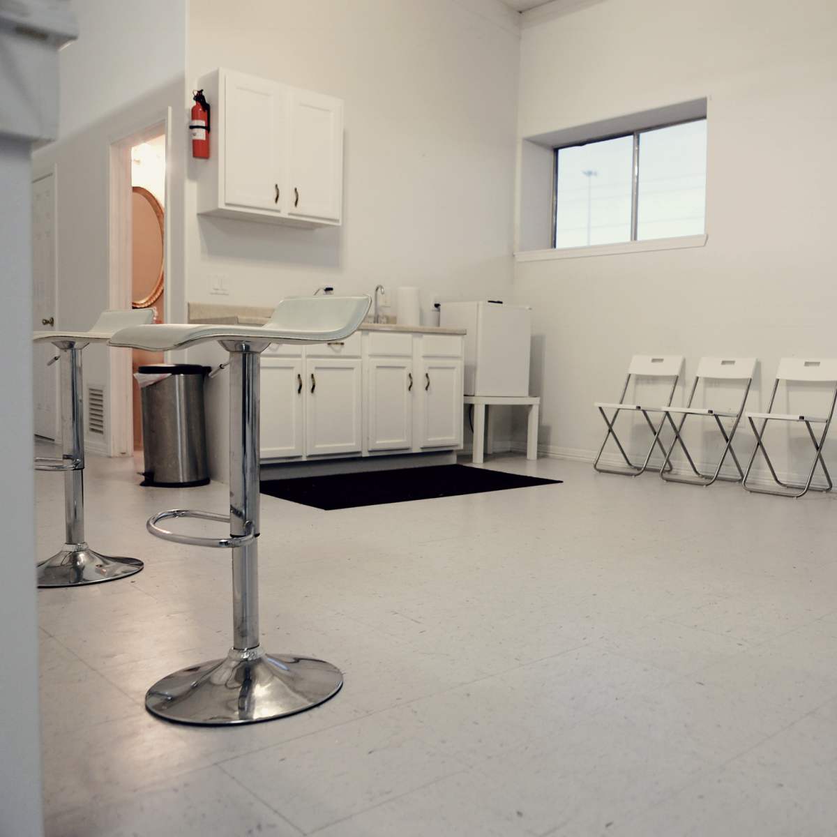 A minimalist waiting room with a row of white folding chairs, a kitchenette area, and a large window letting in natural light.