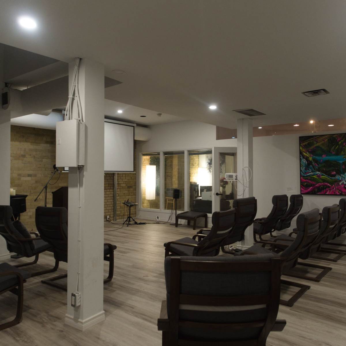 A spacious room with several rows of chairs, a large screen, and a vibrant painting on the wall.