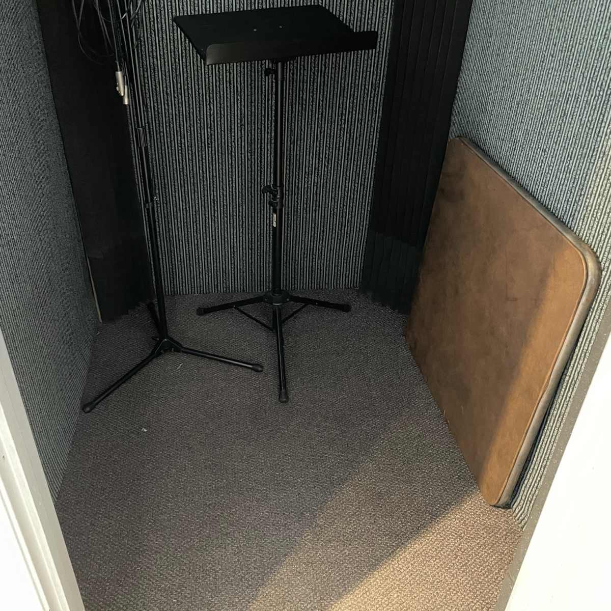 A small soundproof booth with two microphone stands and a folded table against a carpeted floor.