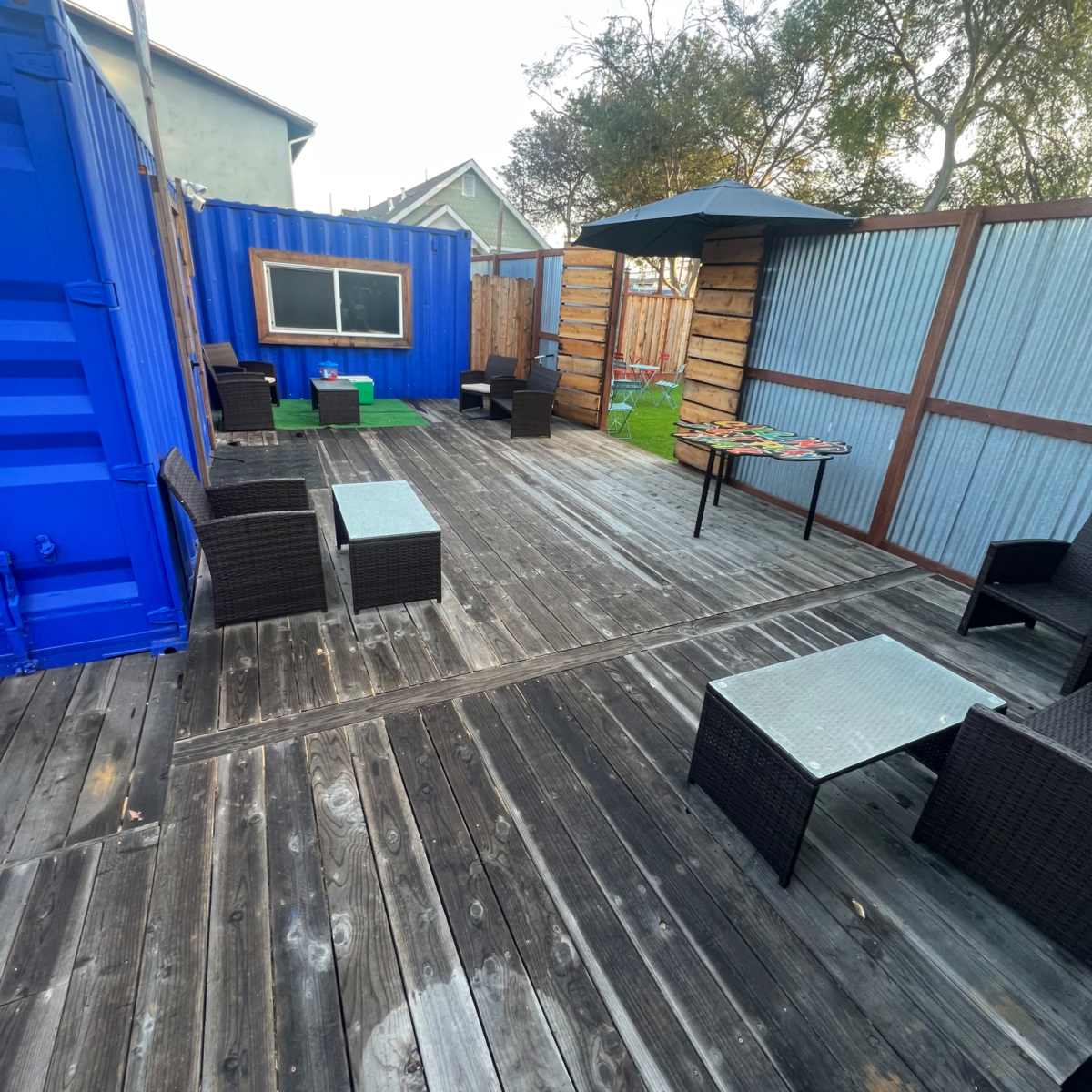 An outdoor seating area with wooden flooring, featuring several wicker chairs and tables, a large blue shipping container, and a small grassy section.