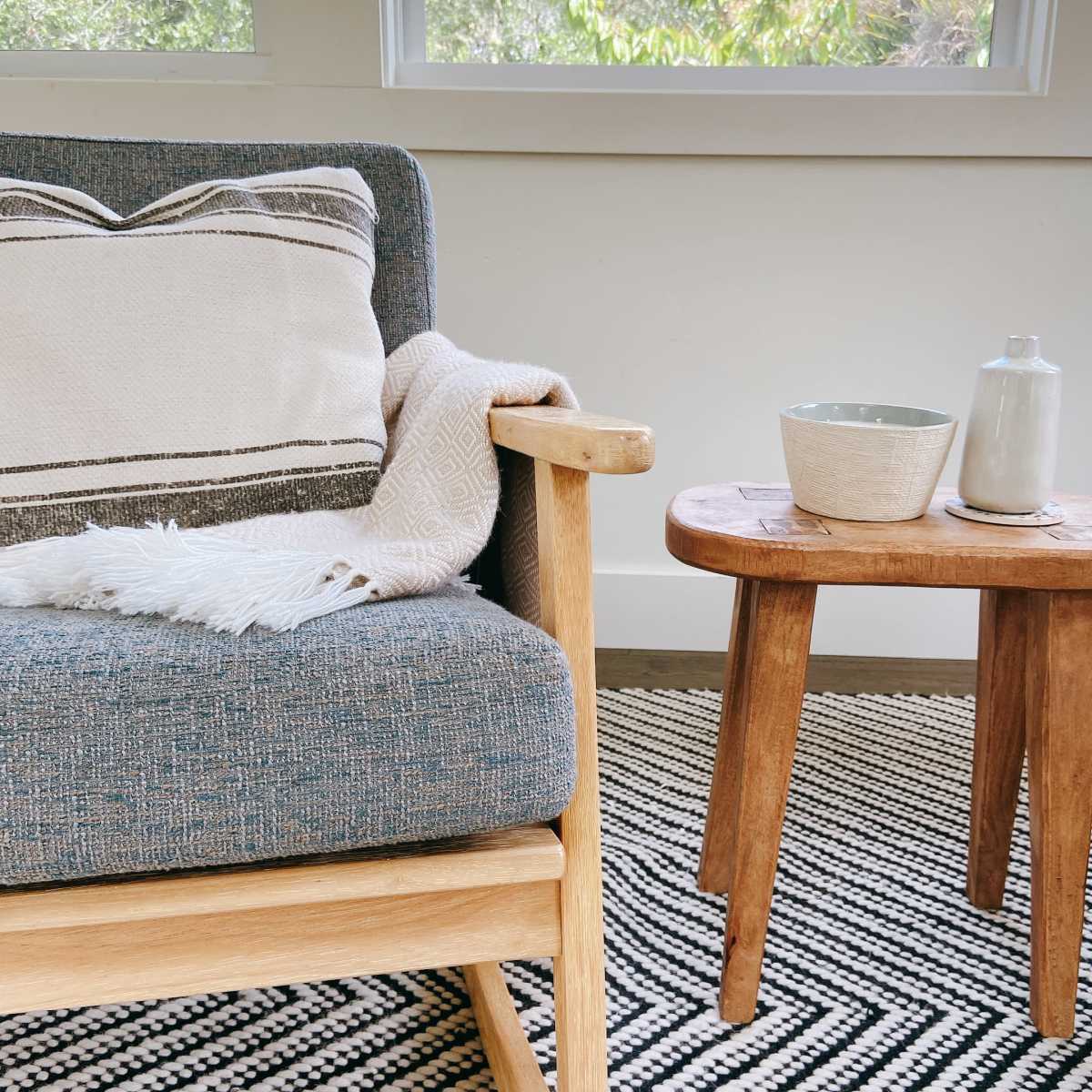 A cozy living room scene features a cushioned chair with a patterned pillow and a throw, alongside a wooden side table with a ceramic bowl and a small pitcher.