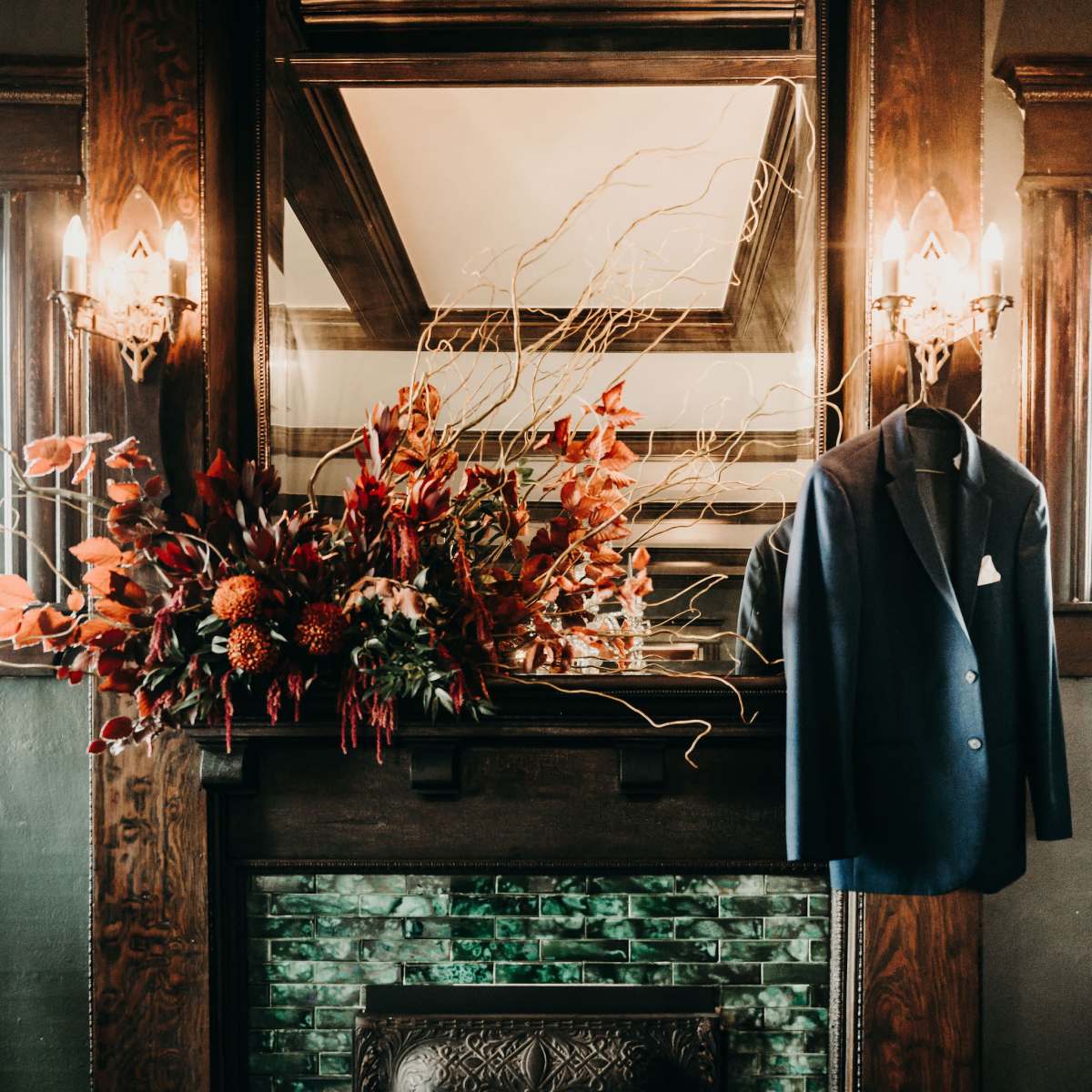 A dark wooden mantel displays an arrangement of autumnal flowers above a vintage green-tiled fireplace, with a navy suit jacket hanging nearby.