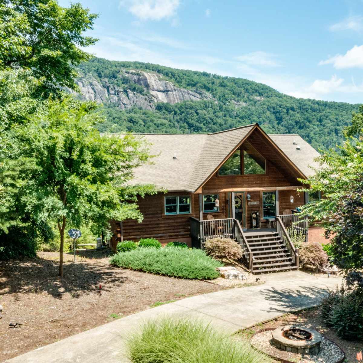 A wooden cabin with large windows and a front porch is situated in a landscaped area, backed by a mountain.