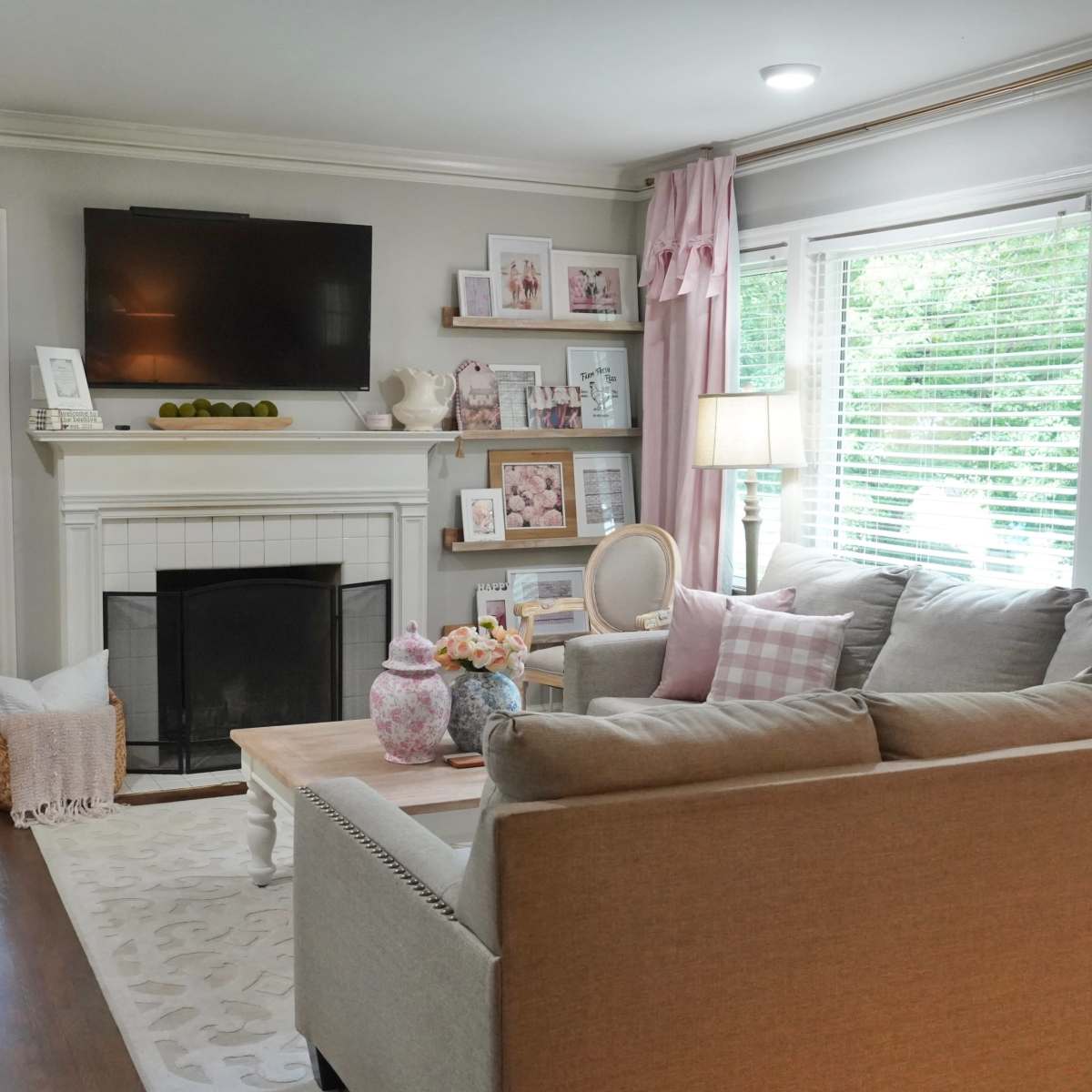 A living room with a light gray couch, wooden coffee table, and a fireplace beneath a mounted television, surrounded by windows with natural light and pink curtains.