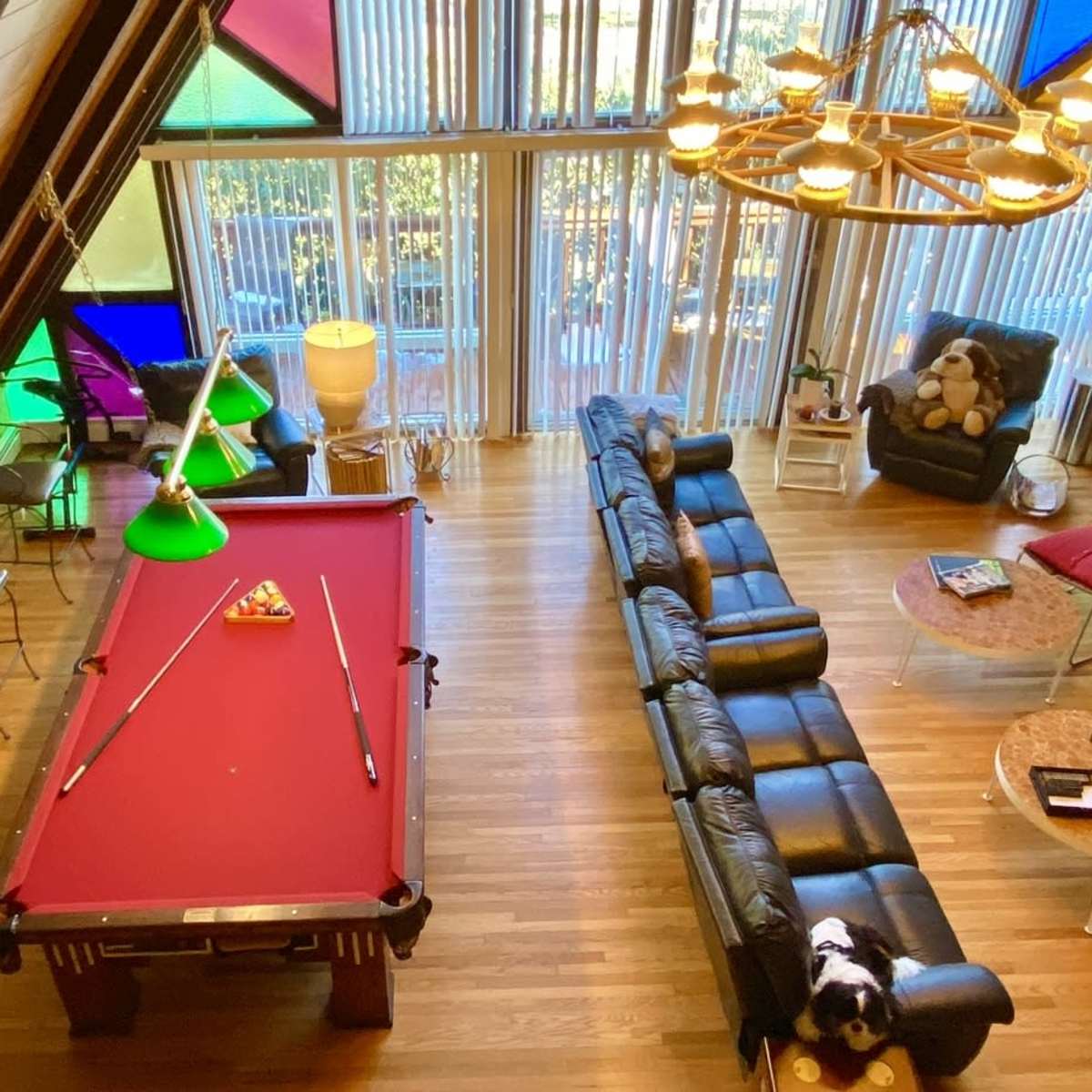 A spacious room with a pool table, a black leather sectional sofa, and large windows featuring colorful glass panels.