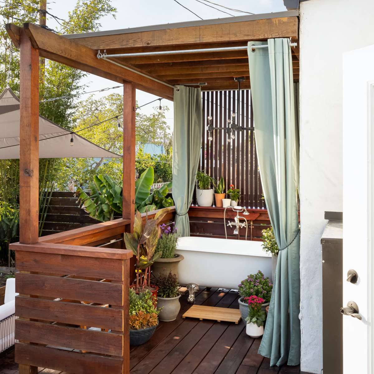 A wooden terrace features a pergola with curtains, surrounded by potted plants and greenery, alongside a building with a white wall.