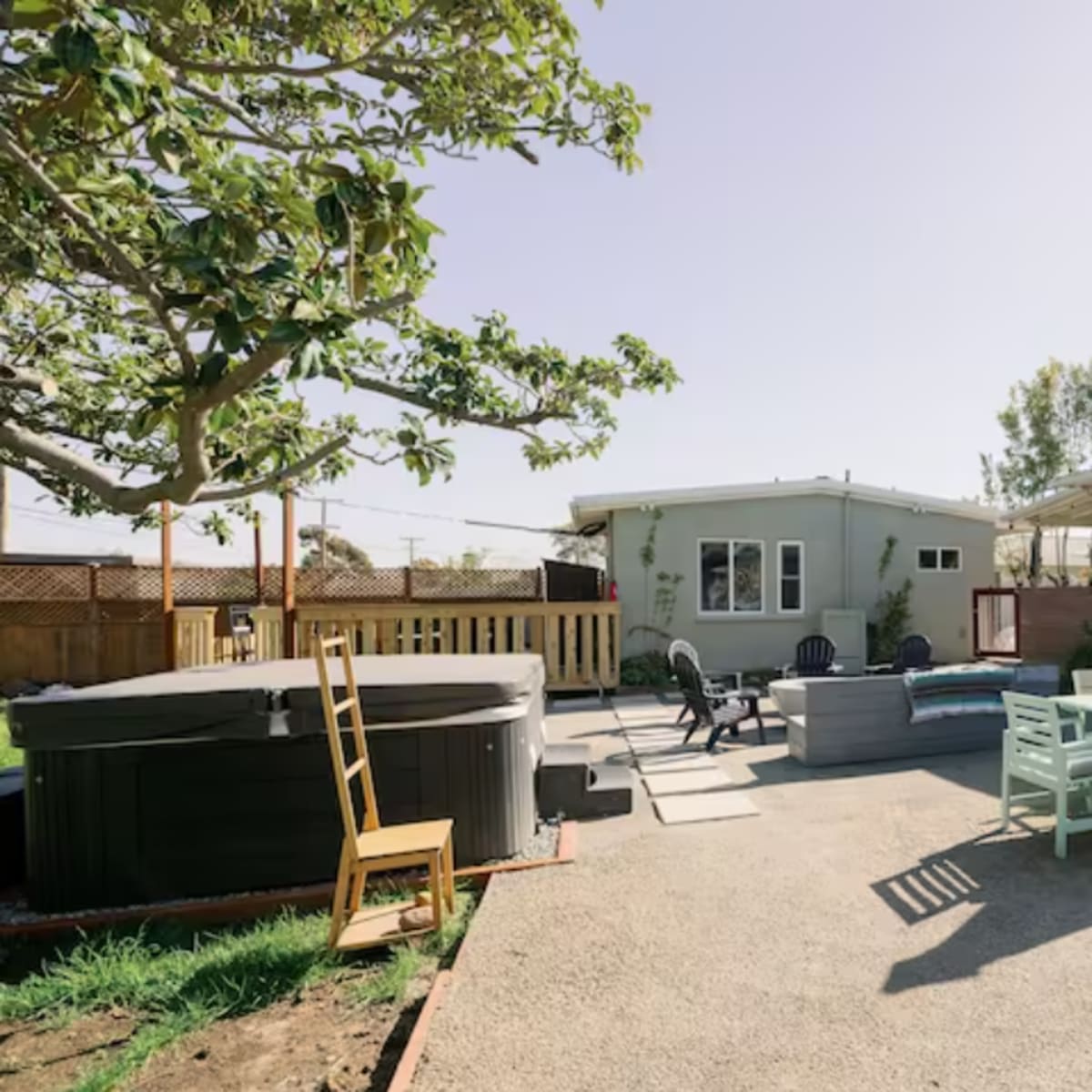 A spacious outdoor area with a hot tub, seating arrangements, and a dining table surrounded by a gravel surface and several trees.