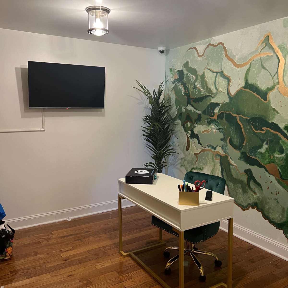 A modern office space featuring a white desk with gold accents, a green chair, a wall decorated with green and gold abstract art, and a flat-screen TV mounted on the wall.