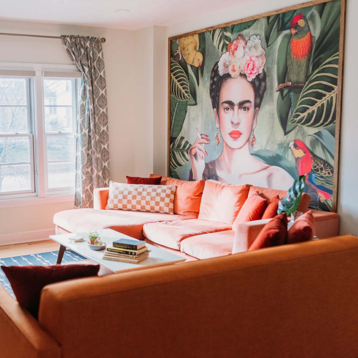 A bright living room features an orange sectional sofa, a large mural of a woman with flowers, and large windows with patterned curtains.