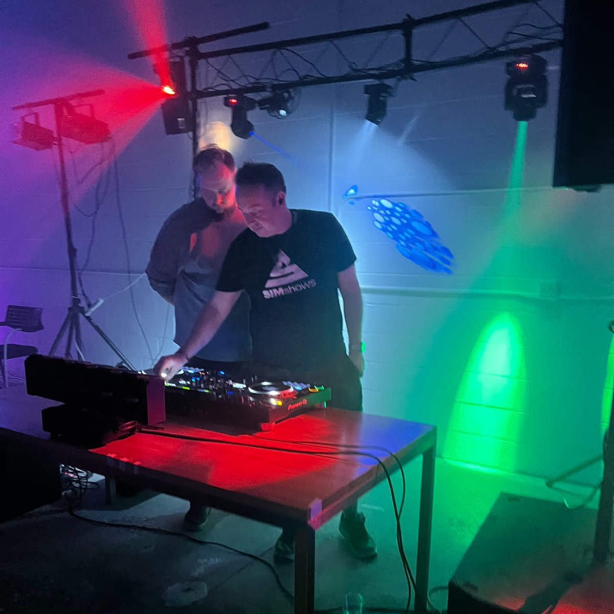 Two DJs operate a mixing console under colorful stage lights in a dimly lit venue.